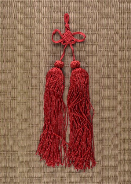 Rote Schwertquaste von Paul Chen. Elegantes, traditionelles Accessoire, das aus feinen, roten Strängen besteht, die kunstvoll in Knoten gebunden sind. Ideal zur Veredelung von Samuraischwertern. Hängt vor einer gewebten Hintergrund.